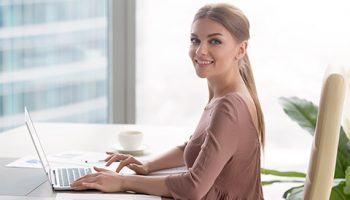 Portrait of smiling young attractive manager, happy businesswoman sitting at workplace using laptop and looking at camera. Starting career in big company, accounting services and business analysis